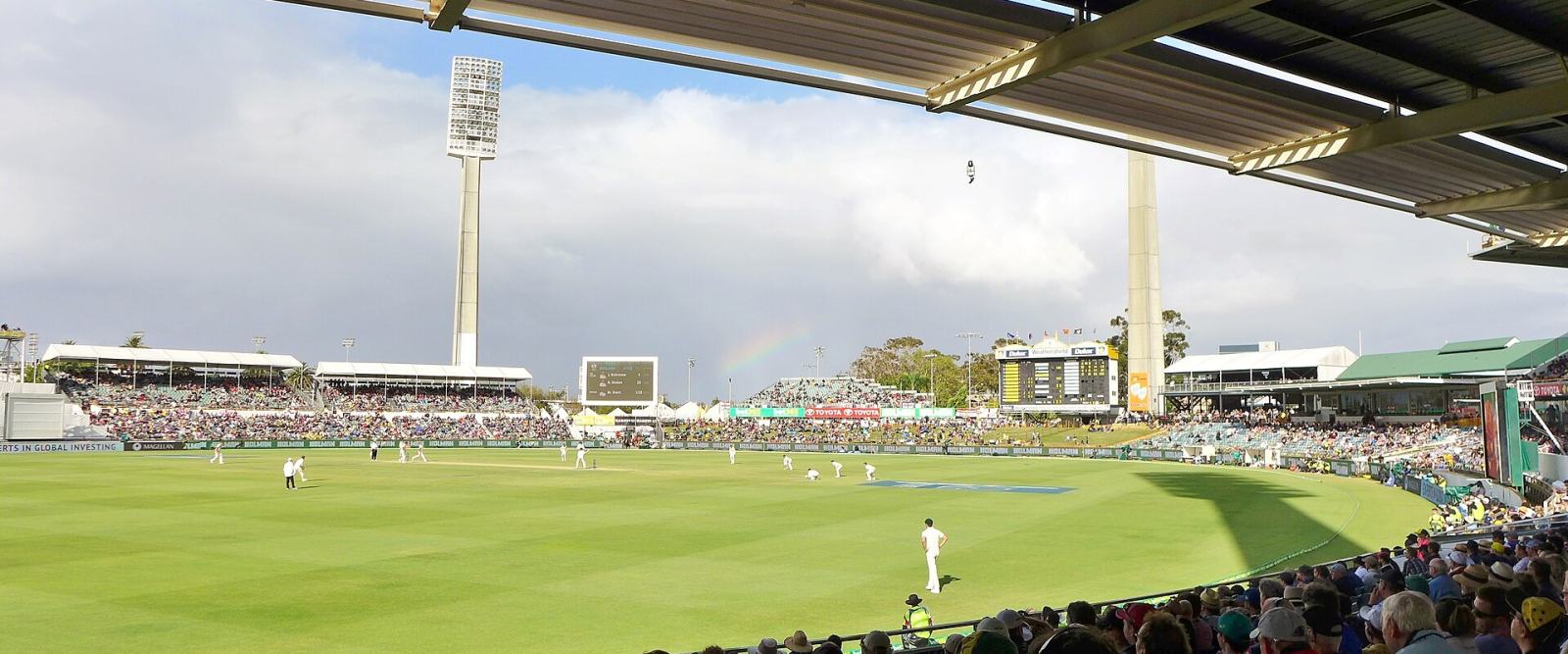 A cricket match in progress on a large Australian ground under a broken sky